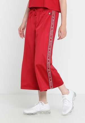 Pantalons larges rouges avec taille élastique à cordon, présentant des rayures latérales avec des détails de logo. Associés à des chaussures de sport blanches.
