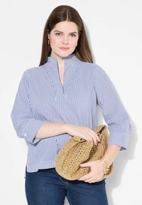 Blusa a righe blu e bianche con colletto alto, maniche 3/4 e dettagli arricciati. Abbinata a una pochette in tessuto dorato e jeans scuri.