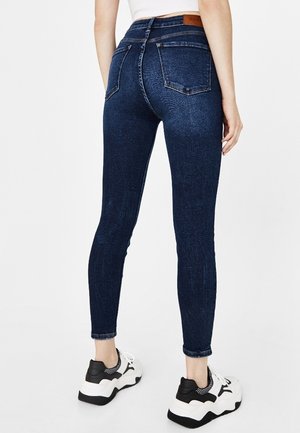 Une personne portant un jean skinny bleu foncé et des baskets blanches et noires, se tenant devant un fond blanc uni.