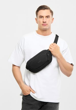 Bărbat purtând un tricou alb și o geantă crossbody neagră, ținând curea cu o mână și sprijinindu-și cealaltă mână în buzunar, pe un fundal simplu.