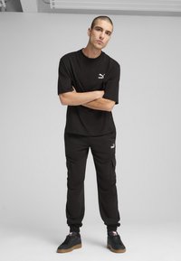 Juoda trumpa rankovė t-shirt su baltu Puma logotipu, derinama su juodais kargo kelnais, turinčiais šonines kišenes ir guma apdailintais padais, dėvima su juodais sportbačiais.