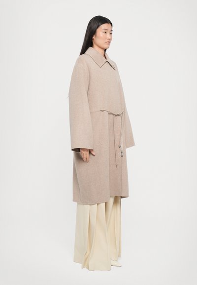Light beige wool coat with a collar, wide sleeves, and a drawstring waist. Worn over light beige wide-legged trousers.