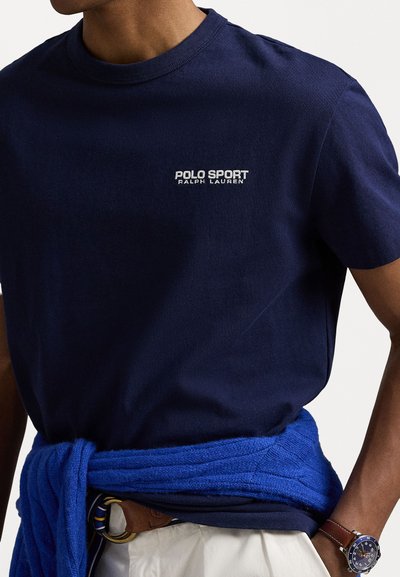 Jūrinės spalvos medvilninis marškinėlis trumpomis rankovėmis, su baltu "POLO SPORT RALPH LAUREN" logotipu krūtinėje. Papildytas mėlyna mezgine džemperiu, surištu aplink liemenį.