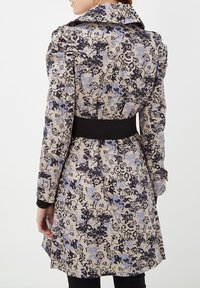 Trench coat de estampado floral con base beige, diseños en azul marino oscuro y azul claro, hombros fruncidos y una cinturilla negra.