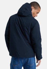 Mørkeblå puffjakke med hætte, der har en glat ydre stof og elastiske manchetter, båret over lyseblå denimjeans.
