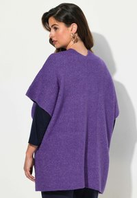 Lila virkad poncho med ribbad textur, med en V-halsringning och korta ärmar, bärs över en svart långärmad topp.