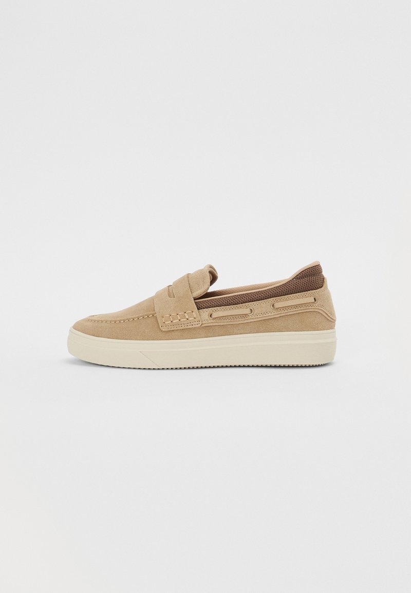 Chaussure beige en daim à enfiler avec des détails cousus, tissu en mesh au talon et semelle épaisse en caoutchouc blanc sur fond uni.
