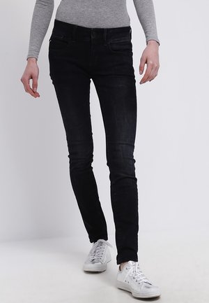 Jeans negros ajustados con un ligero deslavado, diseño de cinco bolsillos y cintura a media altura, combinados con zapatillas blancas y una camiseta gris con relieve.