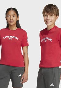 Dvaja mladíci stoja vedľa seba, oblečení v červených tričkách Liverpool FC a sivých šortkách Adidas, na bielom pozadí.