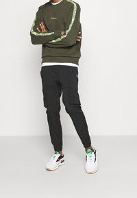 Sudadera verde oliva con detalles de logo en neón, combinada con pantalones cargo negros con bolsillos laterales y zapatillas blancas con acentos de colores.