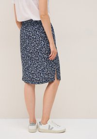 Jupe navy avec un motif de feuilles, présentant une fente sur le côté. Fabriquée en tissu léger, associée à des baskets blanches, montrée sous un angle latéral.