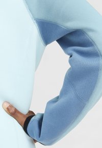 Top atlético azul claro con una manga de color azul oscuro que contrasta. Presenta un tejido suave y elástico, con costuras visibles a lo largo de las uniones.
