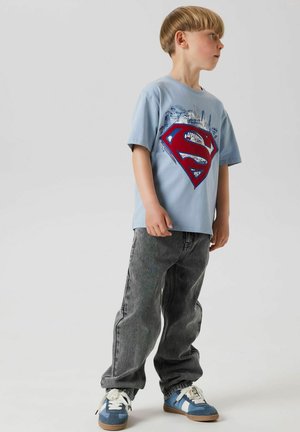 T-shirt bleu clair avec un logo rouge de Superman, accompagné d'un jean baggy gris et de baskets bleues avec des accents blancs. Fond neutre.