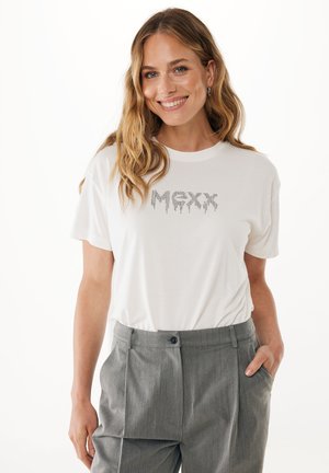 Camiseta de algodón blanca con letras de strass plateadas "MXX" en un diseño goteante. Combinada con pantalones grises de tela plisada.