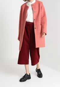 Cappotto di lana rosa con collo rotondo e chiusura a bottoni, abbinato a pantaloni cropped in corduroy rossi e scarpe nere con lacci.