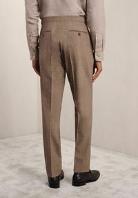 Pantalon beige clair, ajusté, avec une finition texturée, des plis et deux poches arrière, porté avec des mocassins foncés sur une surface douce.