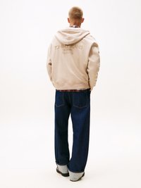 Felpa beige con logo sulla schiena, abbinata a jeans in denim scuro a gamba larga con risvolti. Il tessuto sembra morbido e ha una vestibilità casual.