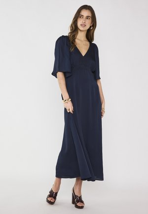 Donna che indossa un abito maxi blu navy con maniche svolazzanti e scollo a v, abbinato a tacchi platform marrone scuro e gioielli in oro.