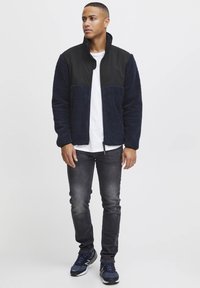 Navy fleece jacket with black nylon panel, zip closure, and side pockets, paired with dark jeans and blue sneakers. Minimal design, casual fit.