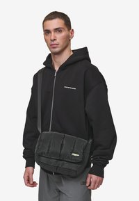 Črna oblazinjena torba za messenger z teksturirano površino, nastavljivim trakom in sprednjim pokrovom. Ima majhen logotip na strani.