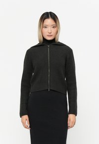 Mørk, teksturert zip-up jakke med hette og lange ermer, med ribbestrikket strikk i nederkant og på mansjettene. Båret over en svart kjole.