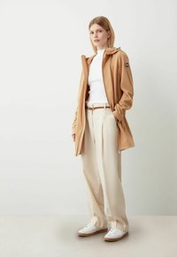 Beige wasserdichte Jacke mit Kapuze, kombiniert mit einem weißen Rippenshirt, beigen Hosen und weißen Sneakers. Auffällige Details sind ein seitlicher Reißverschluss und ein Logo-Patch.