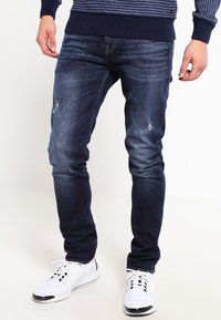 Jeans slim-fit bleu foncé avec une texture délavée et de petites zones usées ; associés à des chaussures de sport blanches et un pull en tricot bleu marine.