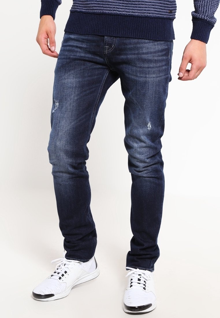 Jeans slim-fit bleu foncé avec une texture délavée et de petites zones usées ; associés à des chaussures de sport blanches et un pull en tricot bleu marine.