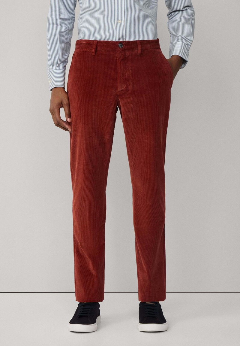 Pantalones de pana en color rojo óxido, con canalones verticales, bolsillos frontales y un corte clásico, combinados con zapatillas oscuras y una camisa a rayas.