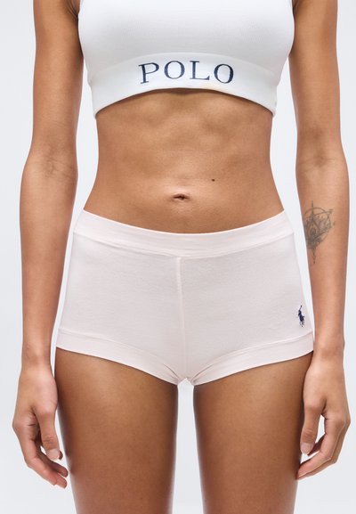 Γυναίκα που φοράει λευκό crop top με το λογότυπο Polo και λευκά εφαρμοστά σορτς με το λογότυπο στο αριστερό μηρό, στέκεται με τα χέρια χαλαρά.
