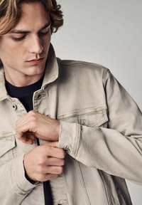 Licht beige denimjas met twee voorzakken en knoopsluiting. Zichtbare stiksels en manchetten op de mouwen.