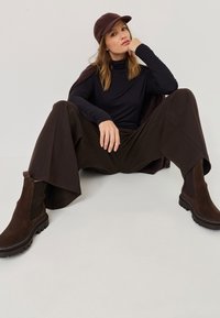 Brązowe zamszowe botki za kostkę z elastycznymi panelami i grubą gumową podeszwą, połączone z ciemnymi szerokimi spodniami i czarnym golfem.