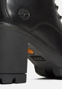 Botte en cuir noir avec talon épais en bloc, logo arbre Timberland embossé sur le côté, et semelle à crampons robuste avec des détails orange et noir.