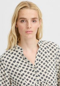 Bluză crem cu model circular bleu marin, fabrică ușoară, placket butonat și design fără guler. Textură netedă și croială lejeră.