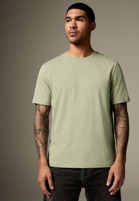 Svetlo zelena bombažna majica z okroglim izrezom, kratkimi rokavi in sproščenim krojem, nošena s temnimi kavbojkami; na rokah so izrezljane črne tetovaže.