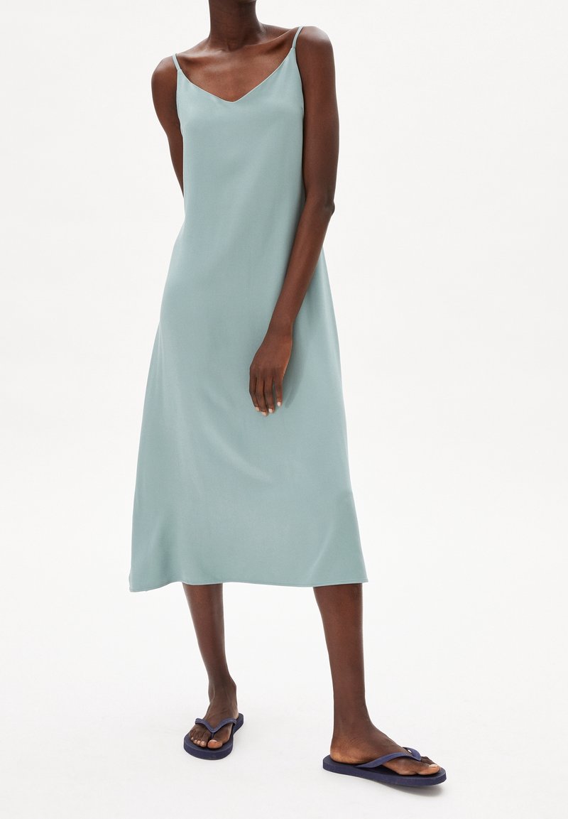 Ein hellgrünes, ärmelloses Midi-Kleid mit V-Ausschnitt, glatter Textur und lockerer Passform, kombiniert mit dunkelblauen Flip-Flops.