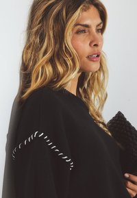 Schwarzer Strickpullover mit weißen gestickten Akzenten an den Ärmeln. Model hält eine schwarze gewebte Clutch, lange gewellte Haare, heller Hintergrund.