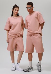 Světlé růžové oversized trička z bavlny a šortky s tkaničkou. Tkaniny jsou měkké a strukturované. Doplňky zahrnují stříbrné šperky a sportovní boty.
