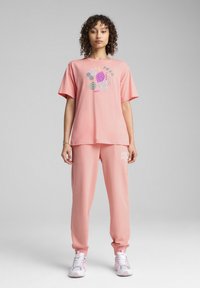 Pink cotton t-shirt with a graphic design and matching pink sweatpants, featuring white logo accents. Model wears light sneakers.