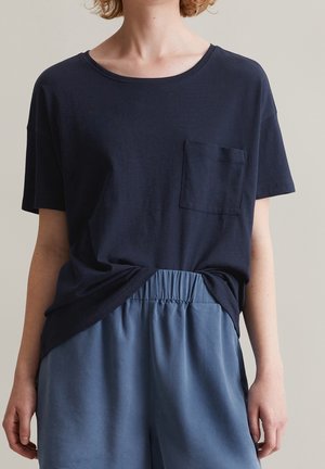 Persoon draagt een marineblauw T-shirt met korte mouwen en borstzak, en losse lichtblauwe broek met elastische tailleband, getoond van schouders tot dijen.
