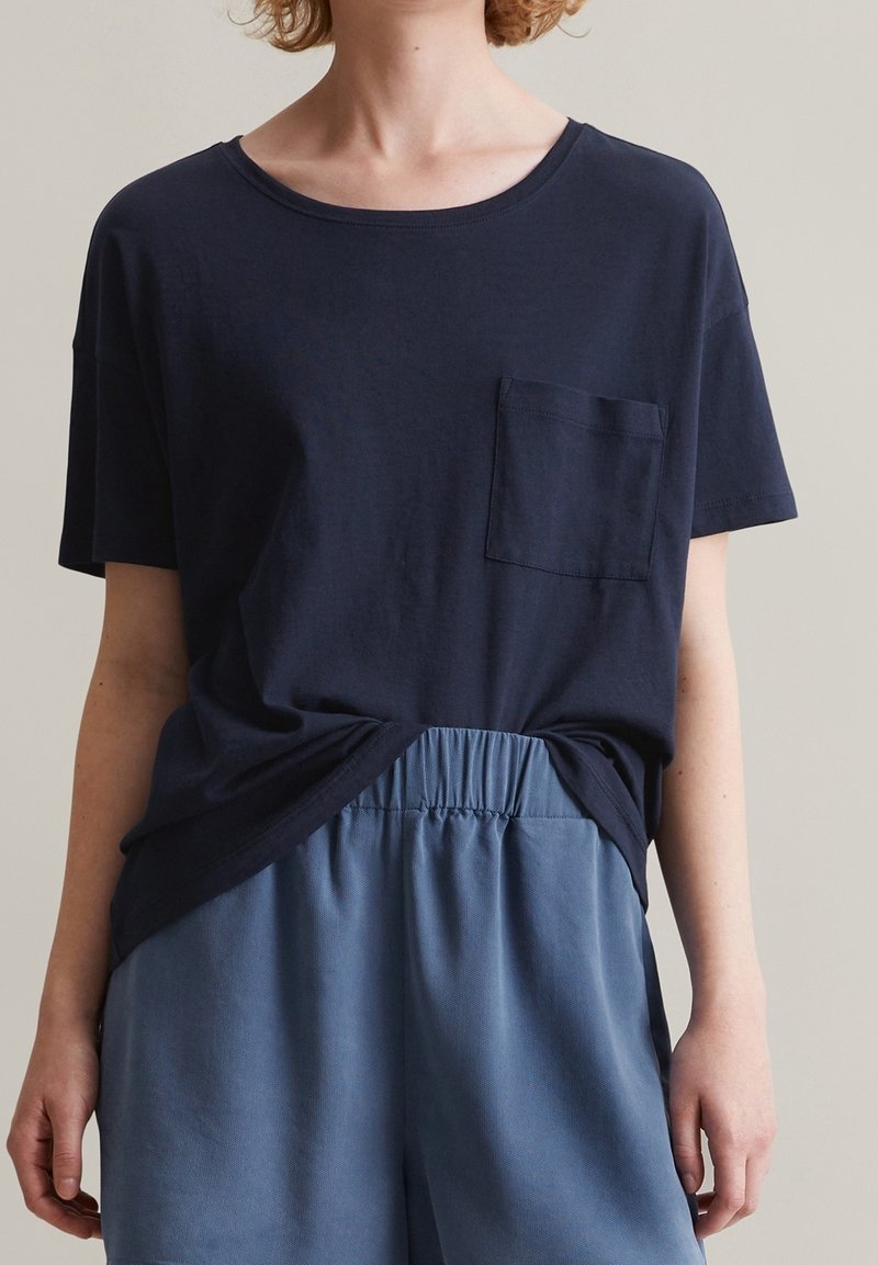 Personne portant un t-shirt bleu marine à manches courtes avec une poche poitrine et un pantalon ample bleu clair à taille élastique, montrée des épaules aux cuisses.
