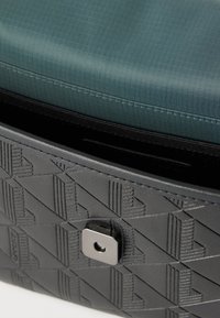 Sac à main en cuir noir embossé avec motif géométrique et fermeture métallique, partiellement ouvert pour révéler une doublure en tissu bleu à l'intérieur.