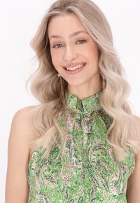 Bluză vernil cu model paisley, cu guler înalt, fabrică ușoară și textură moale. Cercei lungi și delicati adaugă detalii.
