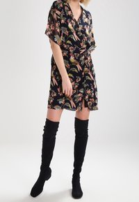 Kwiatowa sukienka w stylu wrap z krótkimi rękawami, głębokim dekoltem w serek, na czarnym tle z różowymi i zielonymi wzorami, zestawiona z czarnymi kozakami do ud.