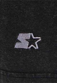 Juodas audinys su tekstūru, kuriame yra pilkai siuvinėti logotipas, sudarytas iš raidės "S" ir žvaigždės formos.