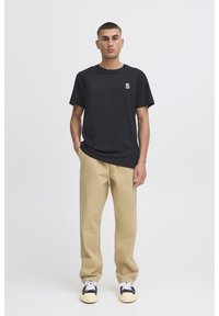 Maglietta di cotone nera con un piccolo logo verde a forma di "S" sul petto, abbinata a pantaloni beige e sneaker bianche e nere.