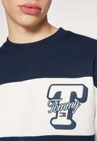 Felpa navy e bianca con colletto a costine, caratterizzata da un grande logo ricamato "T" e "Tommy". Texture liscia, design casual.