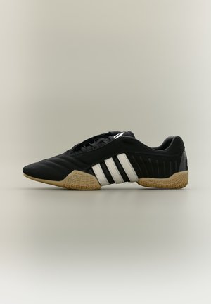 Zapato deportivo negro con rayas blancas y suela texturizada beige sobre un fondo neutro, mostrado de perfil.