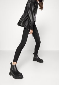 Botas de cuero negras hasta el tobillo con suelas de goma gruesas, paneles elásticos laterales y costuras en contraste. Combinadas con jeans negros ajustados y una chaqueta negra.