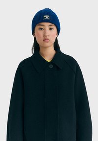 Joven con un gorro de punto azul con un parche y un abrigo negro abotonado sobre una camiseta amarilla, mirando hacia adelante con una expresión neutral.